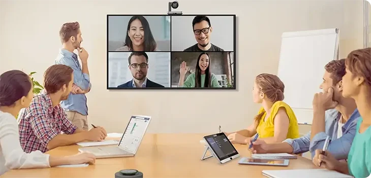 Six colleagues in a conference room, engaged in a video call with four people displayed on a large screen at the front.