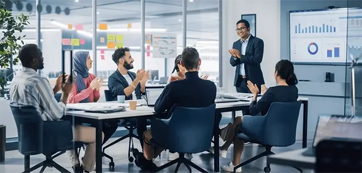 A diverse group of colleagues in a modern office applauding a standing man, with charts displayed on a screen in the background.